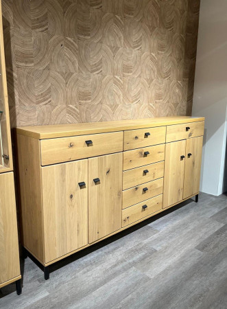 Sideboard Vilano Solero aus heller Asche-Massivholz mit mattschwarzen Griffen vor einer hellbraunen Wand und grauem Holzboden.