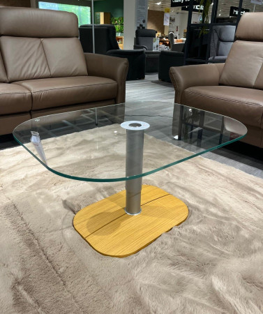 Couchtisch Düren mit Glasplatte, Eichenfurnier und Metallgestell auf einem beigen Teppich, zwischen braunen Ledersofas im Ausstellungsraum.