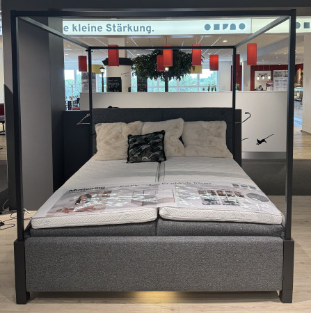 Ein modernes Himmelbett „Musterring MyHeaven“ (180 x 200) mit grauem Bezug und flauschigen Kissen in einem Ausstellungsraum.