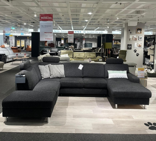 Dunkelgraues Ecksofa Planoform Nizza mit Federkern, Stoff Vincent, abgebildet mit Kissen in einer Ausstellungsumgebung.
