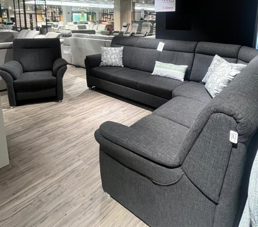 Wohnlandschaft „Planoform Favorit“ in Dunkelgrau mit Sessel, präsentiert auf einem Holzboden in einem modernen Ausstellungsraum.