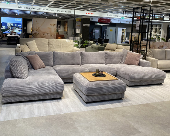 Wohnlandschaft „Hagerstown“: Großes graues Sofa mit Rückenkissen, Ottomane und rosa Kissen im Ausstellungsraum.