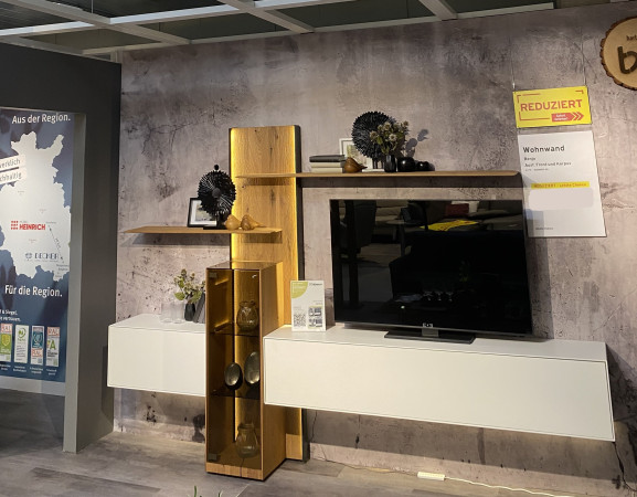Wohnwand „Benjo“: Moderne Wohnwand mit Regalen, Fernseher, goldenen Akzenten und Riffholz-Details in einem stilvollen Ausstellungsraum.