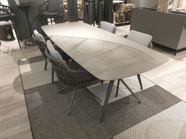 Tischgruppe „Cliff/Andria“: moderner Tisch aus grauem Geflecht, 6 Stühle mit Olefin-Kissen, auf einem gemusterten Teppich.