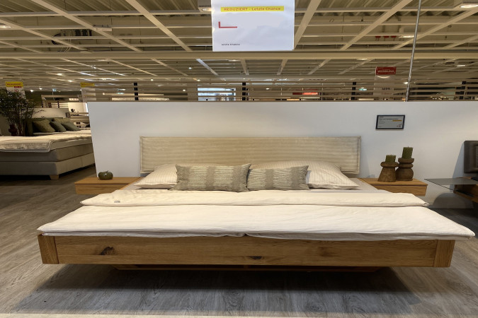 Das Bett mit Nachttischen (Freiburg 11) mit beigem Kopfteil wird in einem modernen Möbelausstellungsraum präsentiert.
