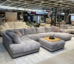 Wohnlandschaft „Hagerstown“ in Grau mit Hocker und Holztablett, ausgestellt im modernen Showroom neben weiteren Sofas.