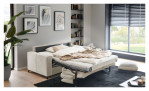 Das Komfort-Schlafsofa „Pickering“ von Hukla in einem modernen Wohnzimmer mit Wanddekoration, Bücherregalen und einem großen Fenster.