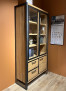 Vitrine „Metalo“ – hoher Holzschrank mit Glastüren, Metallrahmen, Einlegeböden, 4 Schubladen, Wandfarbe „Railway Brown“.