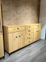 Sideboard Vilano Solero aus heller Asche-Massivholz mit mattschwarzen Griffen vor einer hellbraunen Wand und grauem Holzboden.