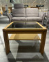 Couchtisch Venta Plus aus massiver Wildeiche mit Glasplatte und Ablage, im Geschäft vor einem grauen Sofa ausgestellt.
