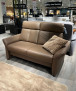 Sitzgruppe Mondo Comfort Evado aus braunem Leder mit beigem Kissen, abgebildet in einem Ausstellungsraum neben anderen Sofas.