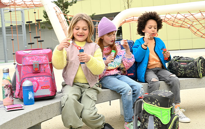 Drei Kinder sitzen auf einer Bank und machen Seifenblasen, neben ihnen bunte Schulranzen und Trinkflaschen