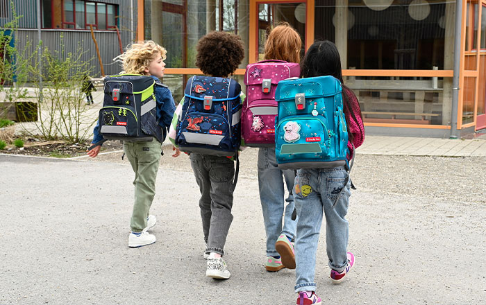 Drei Kinder mit bunten Schulranzen gehen auf eine Schule zu