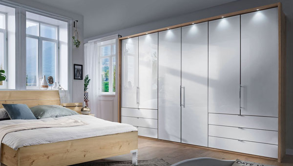 Komfort-Kleiderschrank Rügen mit mehreren Türen, Schubladen und integrierter Beleuchtung neben einem Holzbet