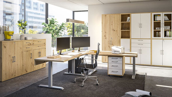 Modernes Büro mit Eckschreibtisch aus hellem Holz, zwei Monitoren, Schreibtischstuhl und großen Schränken mit weißen Fronten