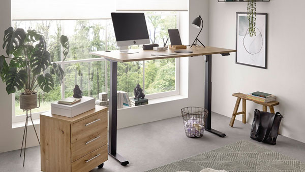 Höhenverstellbarer Schreibtisch mit Holzplatte und schwarzen Metallbeinen, darauf Computer und Lampe, daneben Rollcontainer