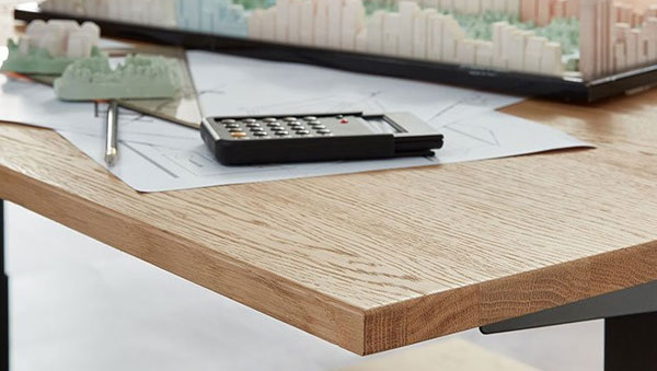 Eckansicht eines Schreibtischs mit Holzfurnier, darauf liegen ein Taschenrechner, Baupläne und ein Modell einer Stadt