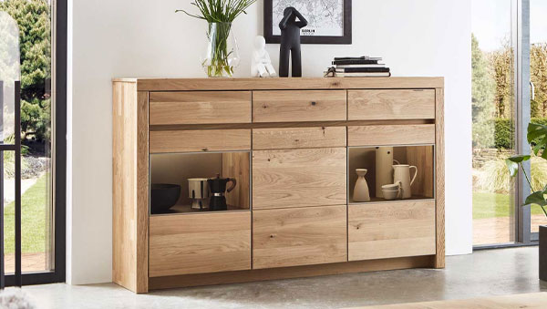 Basis-Highboard witlake Soraga aus Massivholz mit Schubladen, Türen und zwei offenen Fächern, dekoriert mit Geschirr, Büchern und Vasen vor großen Fenstern