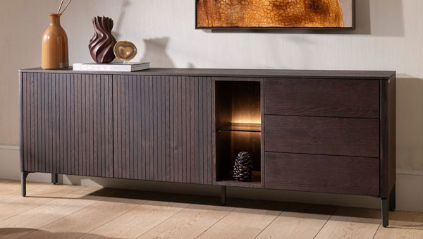 Sideboard Trenton mit geriffelter Front, Schubladen, beleuchtetem offenen Fach und schlanken Metallfüßen vor einer hellen Wand