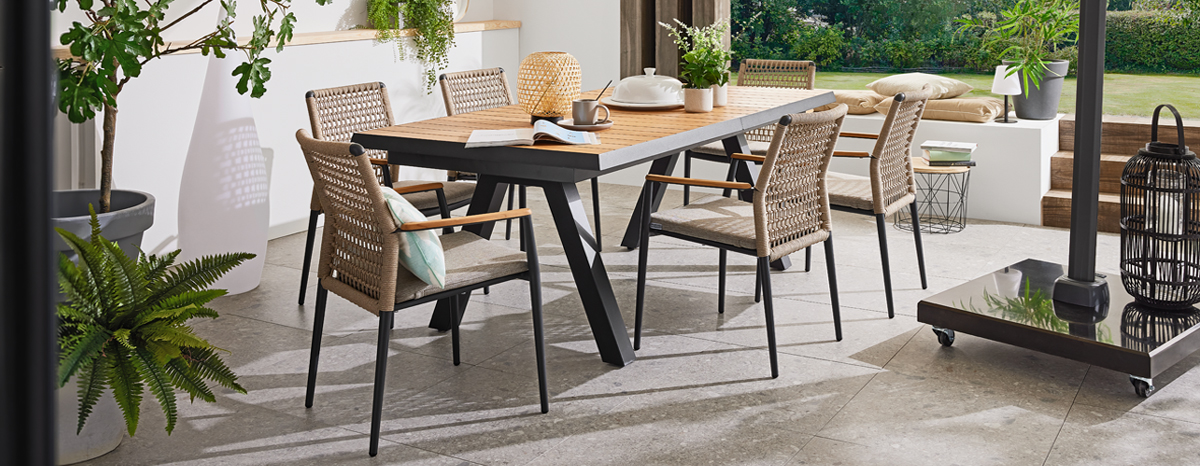 Gartenmöbel MONDO Latea mit Teakholz-Gartentisch und sechs stapelbaren Stühlen mit Rope-Geflecht, Sitzkissen in Taupe und Teakholz-Armlehnen auf Terrasse mit Gartenblick