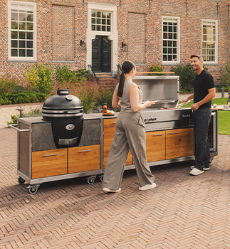 Zwei Personen stehen an einer Outdoorküche mit Grill Pro Master und Kamado Grillmodul vor einem Backsteingebäude