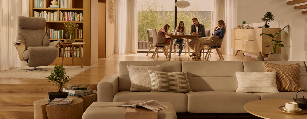 Modernes Wohnzimmer mit Stressless Möbeln: beige Stressless Couch mit zwei Kissen, Stressless Sessel und Holzmöbeln; im Hintergrund sitzen vier Personen am Esstisch.