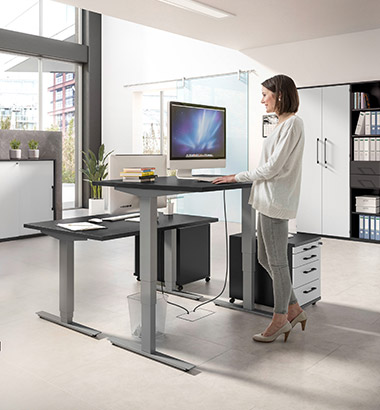 Frau steht an Röhr Teamarbeitsplatz mit höhenverstellbaren Schreibtischen in Grau mit schwarzer Tischplatte
